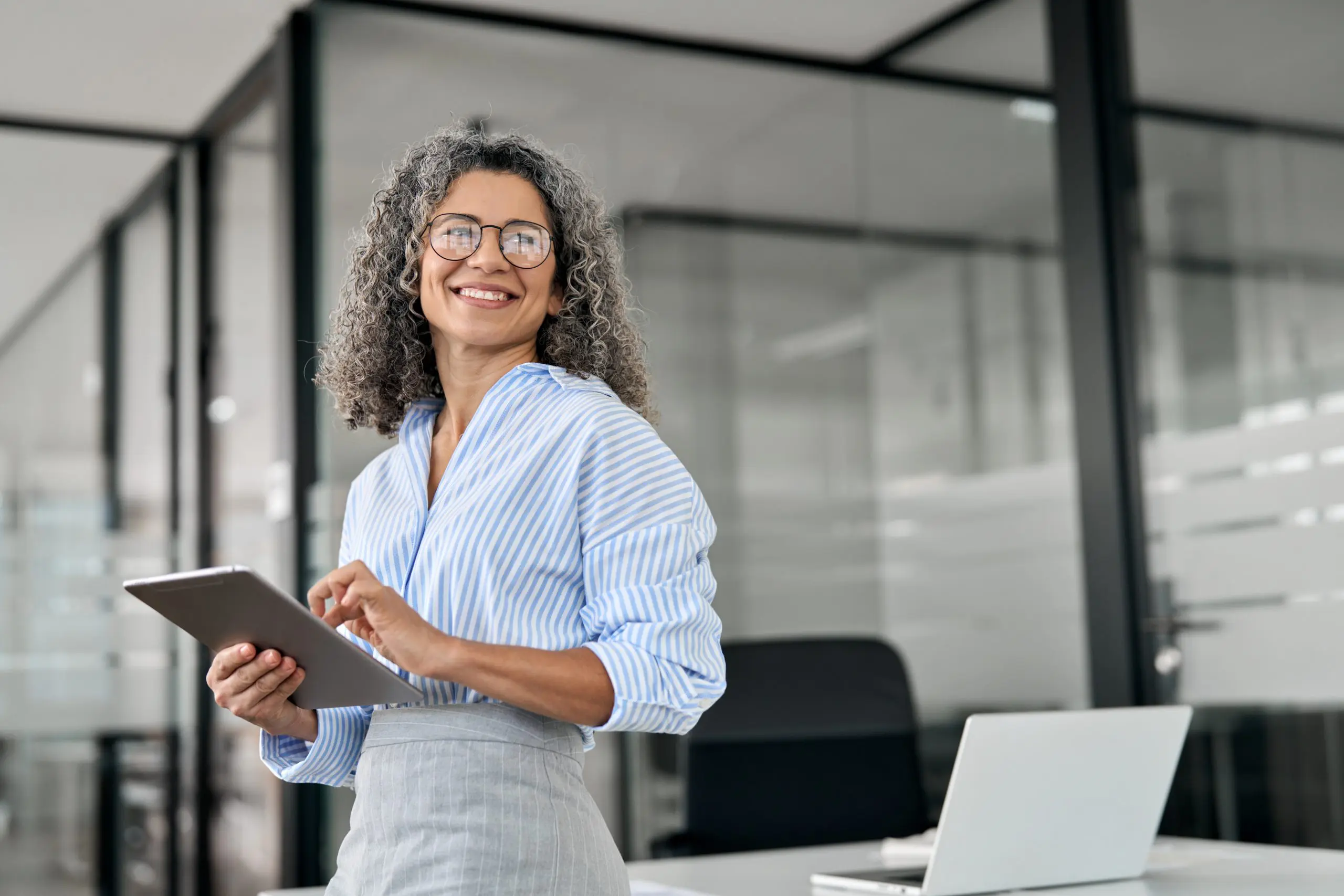 Smiling professional woman with curly gray hair, wearing glasses and a blue striped shirt, standing in a modern office while holding a tablet. A laptop is visible on the desk behind her.