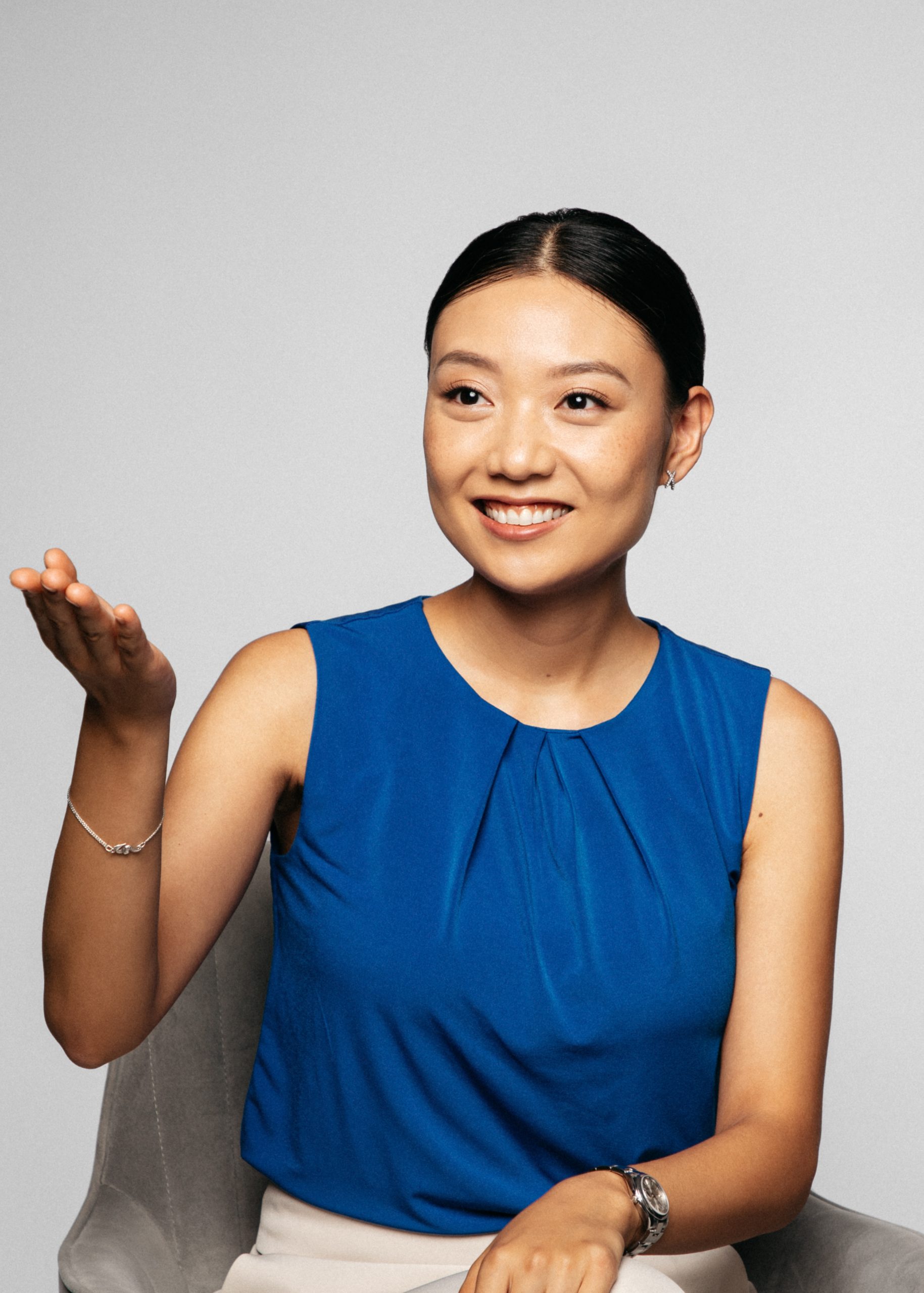 Smiling woman in a blue sleeveless top gesturing with her hand, against a neutral background, showcasing confidence and approachability.