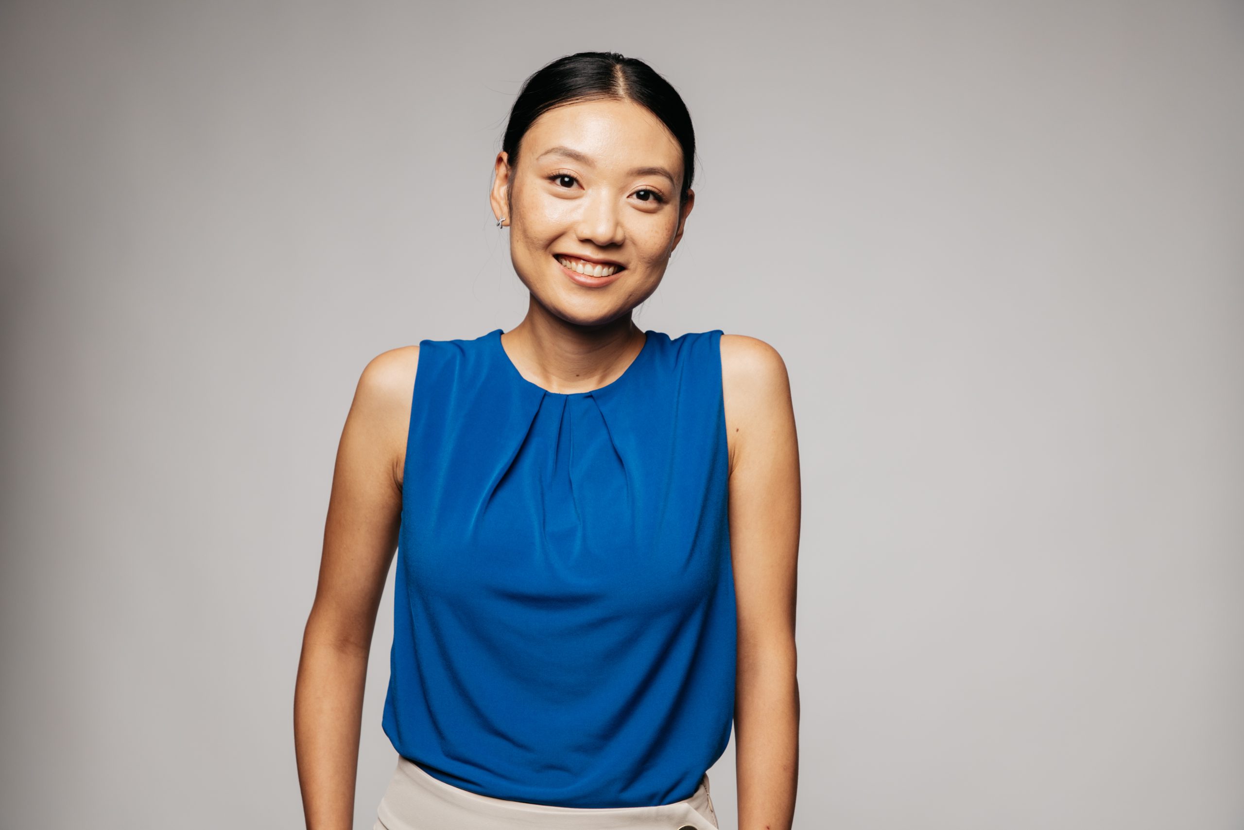 Smiling young woman in a blue blouse, standing against a neutral background, showcasing a friendly and approachable demeanor.