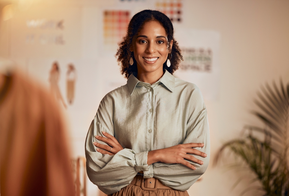Smiling woman with curly hair wearing a light green shirt and beige pants, standing with arms crossed in a creative workspace with colorful design samples in the background.