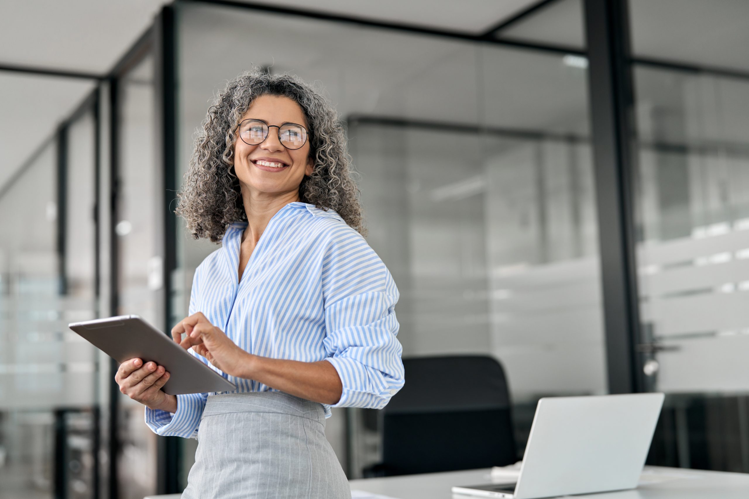 Smiling professional woman with curly gray hair, wearing glasses and a blue striped shirt, standing in a modern office while holding a tablet. A laptop is visible on the desk behind her.