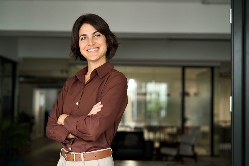 Smiling woman with short dark hair standing confidently with arms crossed in a modern office setting.