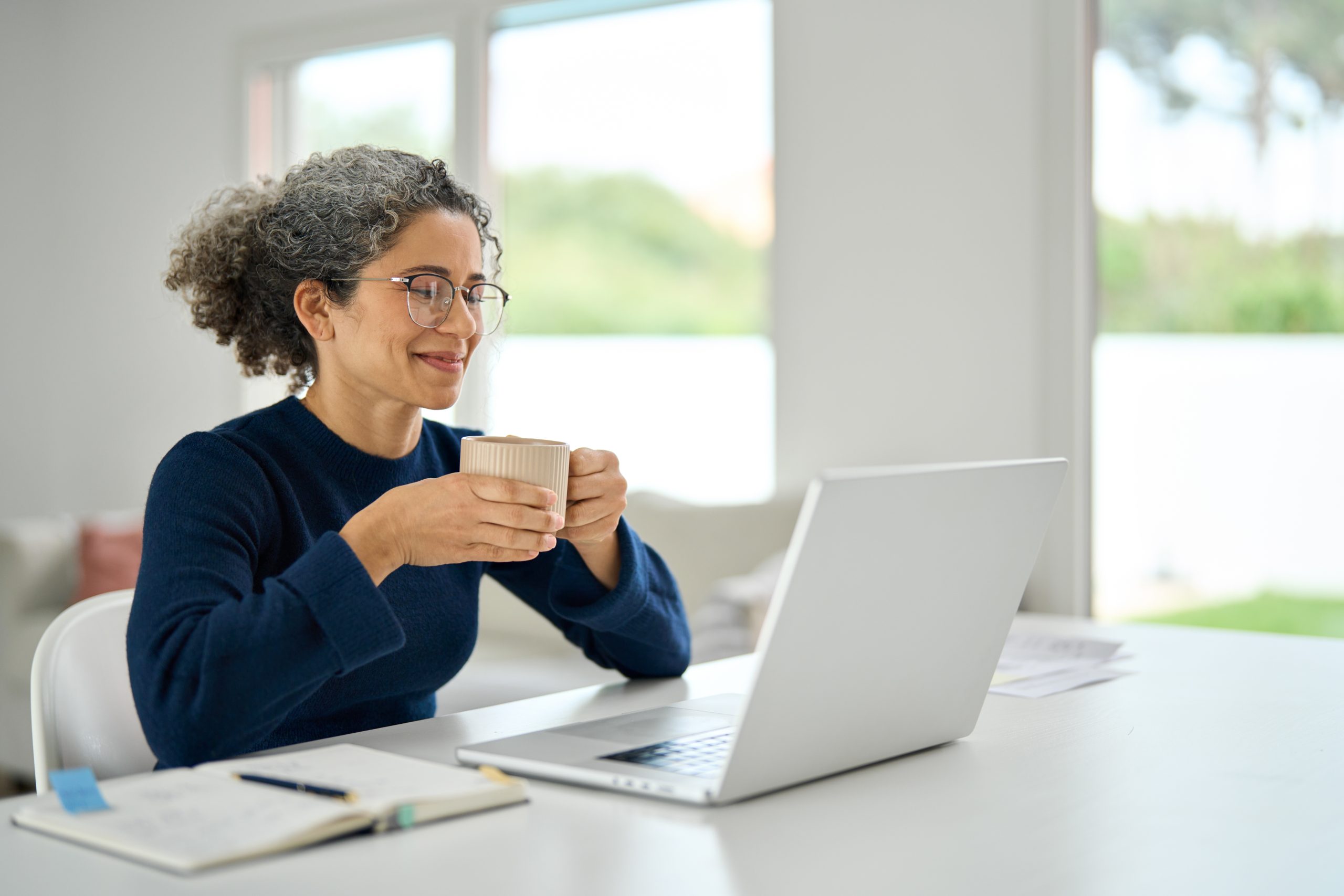 Woman enjoying a warm drink while working on a laptop at a bright, modern home office. She is wearing glasses and a navy sweater, with a notebook and pen on the table.