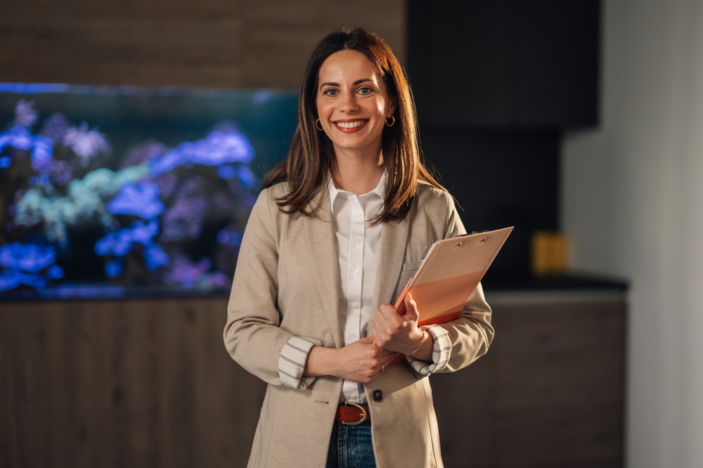 Smiling professional woman holding a folder, dressed in a beige coat and white shirt, standing in a modern office with an aquarium in the background.