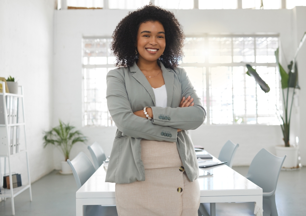 Confident businesswoman with curly hair smiling in a modern office setting, wearing a light gray blazer and beige skirt, standing with arms crossed.