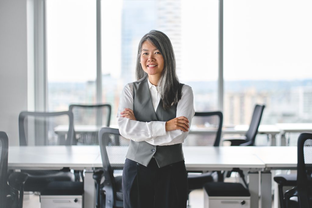 Professional woman standing confidently in a modern office environment, wearing a gray vest over a white shirt, with arms crossed and a smile. Bright windows in the background showcase a city view, emphasizing a corporate atmosphere.