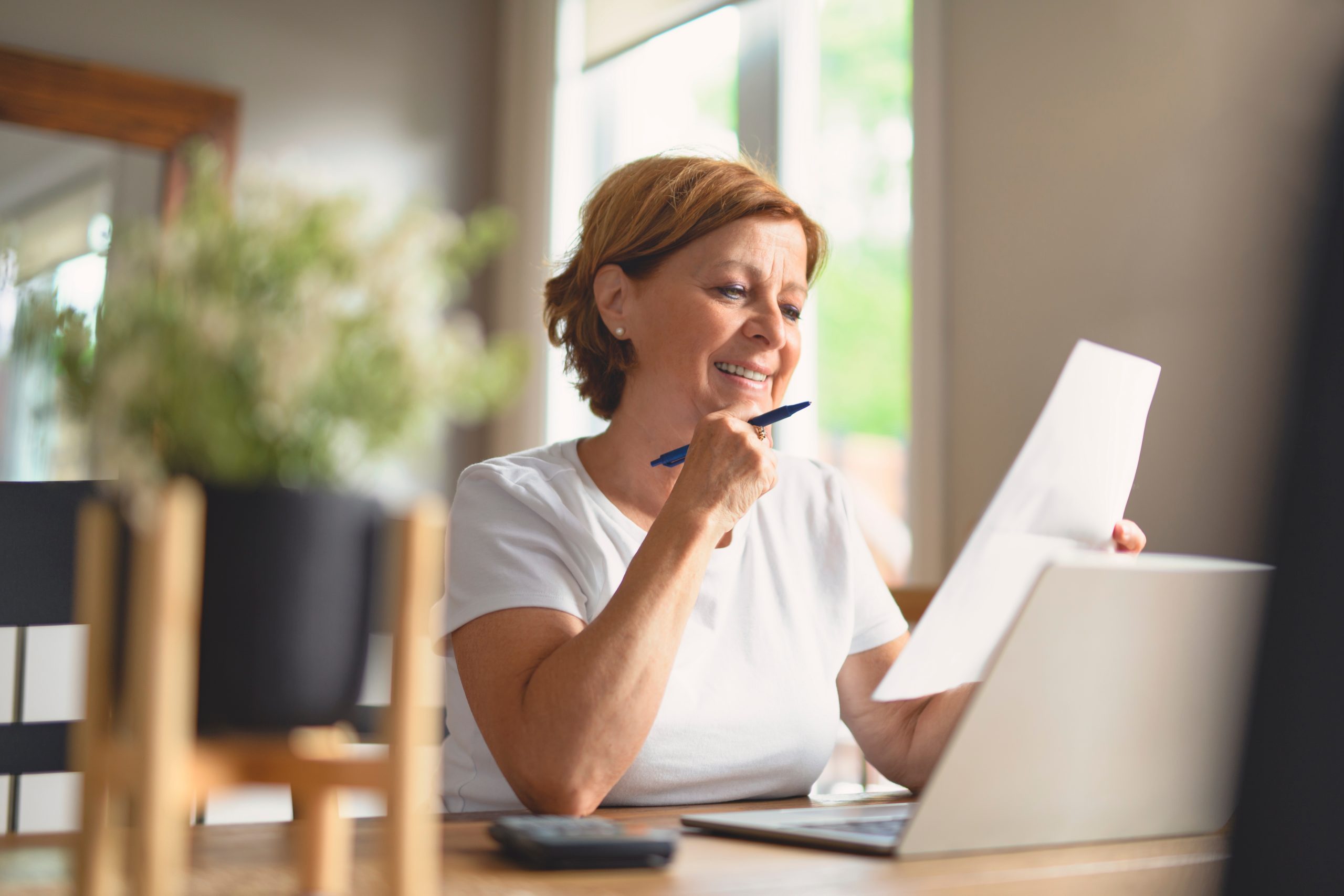 Smiling woman sitting at a wooden table, reviewing documents while using a laptop, with a pen in hand and a potted plant in the background.
