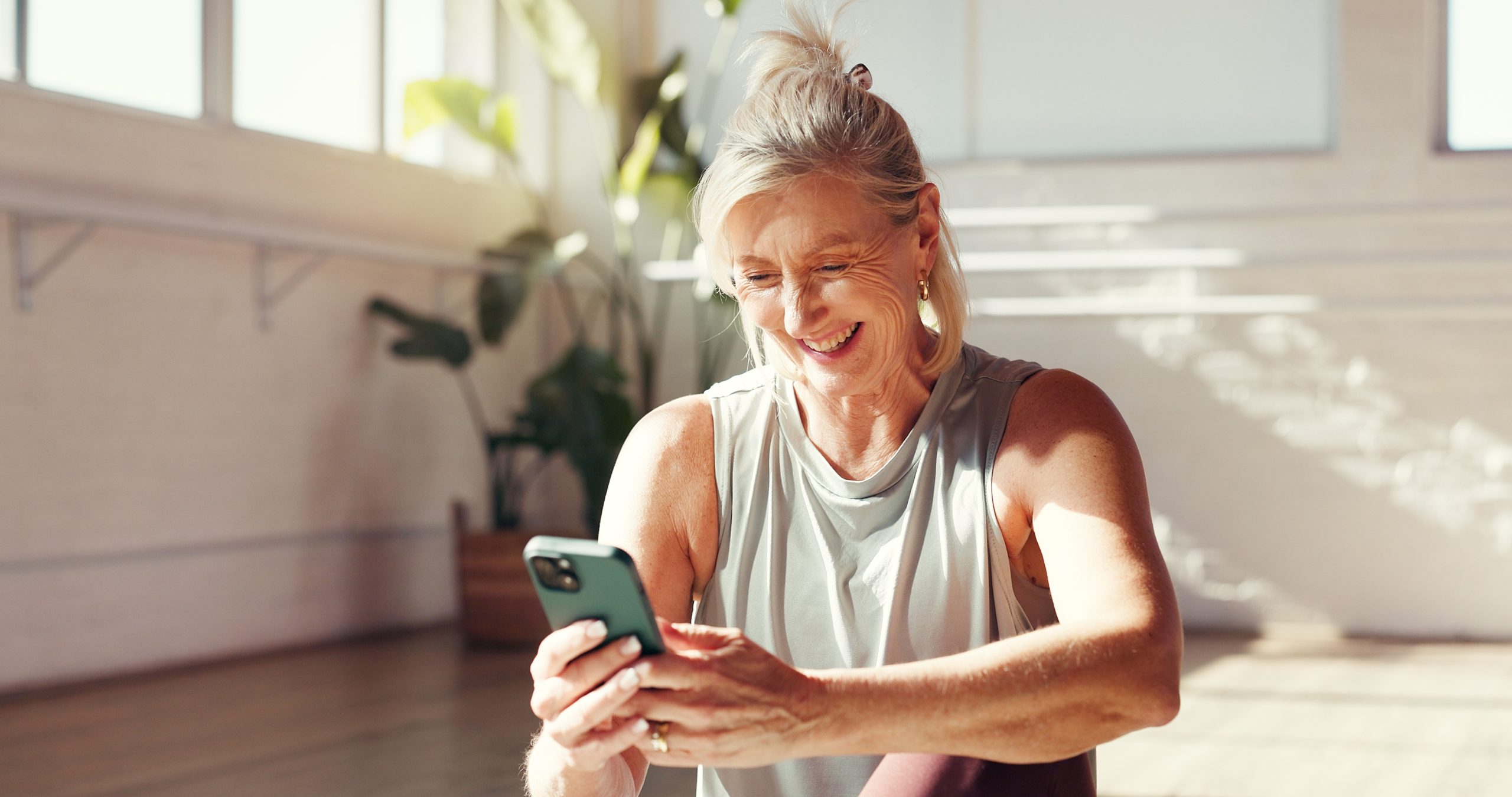 Smiling older woman in workout attire using a smartphone while sitting in a bright, airy room with plants, promoting a healthy and active lifestyle.