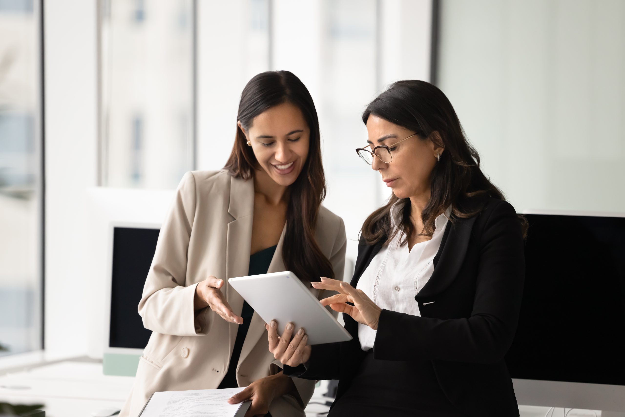 Two businesswomen collaborating in a modern office, discussing a project on a tablet. One woman is wearing a beige blazer and the other a black suit, both engaged in a productive conversation.