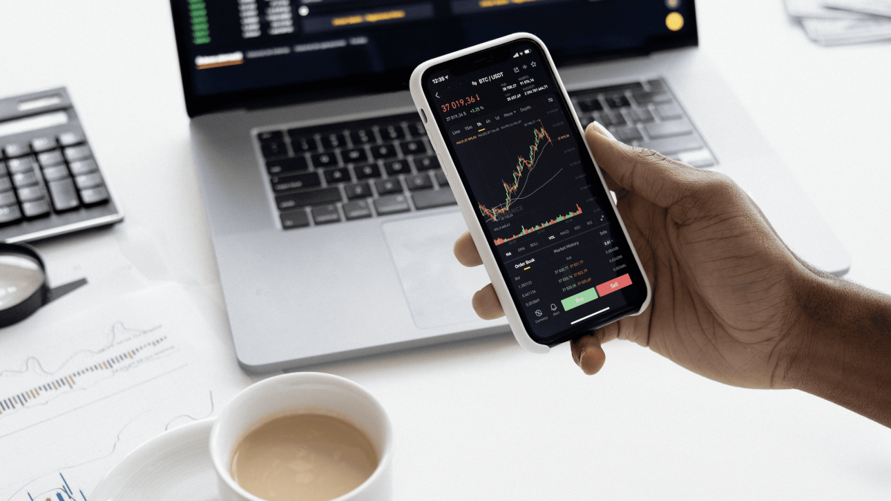 stock market on the laptop and on a mobile device and a cup of coffee and calculator next to them