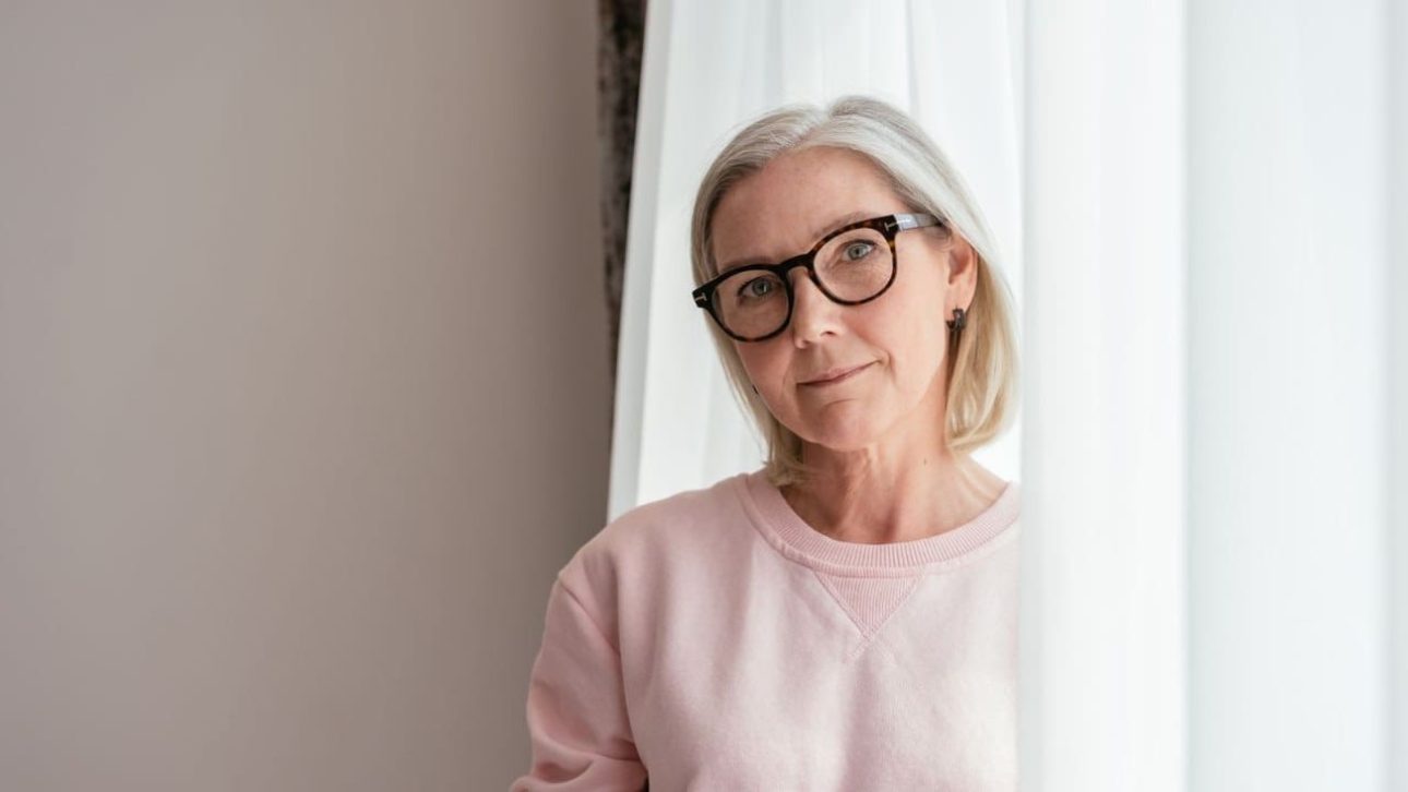 Composed woman in her 60s standing by a bright window, thinking through financial decisions after loss