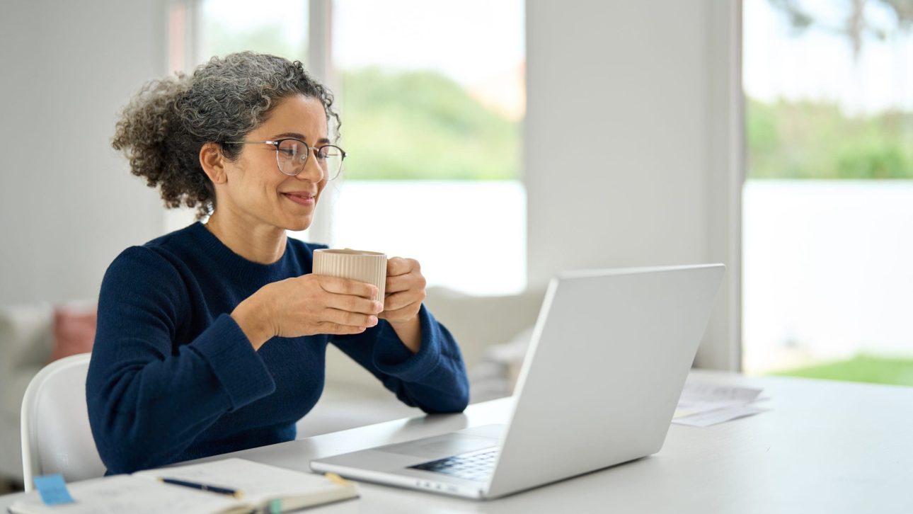 Woman enjoying a warm drink while working on a laptop at a bright, modern home office. She is wearing glasses and a navy sweater, with a notebook and pen on the table.